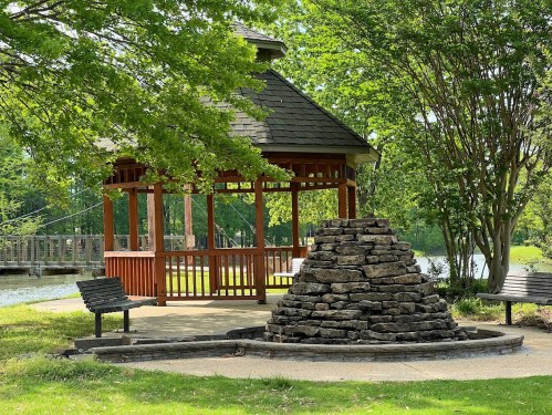 A gazebo surrounded by greenery, with a stone structure and benches nearby, beside a calm body of water.