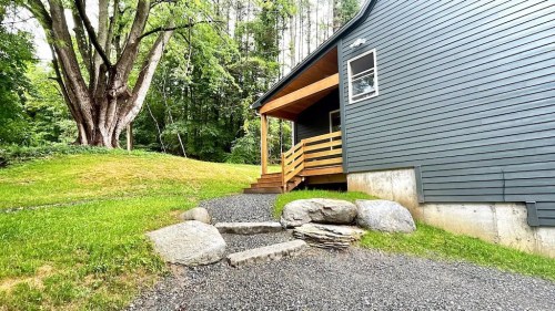 A pathway of stones leads to a modern gray house with a wooden porch, surrounded by lush greenery and trees.