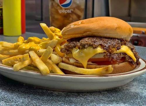 A juicy cheeseburger with melted cheese, tomato, and fries drizzled with mustard on a plate, with a soda in the background.