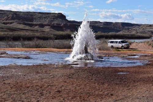 A geyser erupts in a dry landscape, with a vehicle parked nearby and rocky hills in the background under a blue sky.