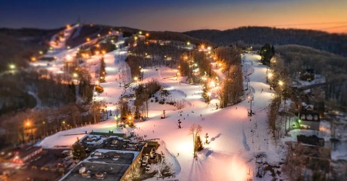 Aerial view of a ski resort at dusk, with illuminated slopes and skiers on snowy trails surrounded by trees.