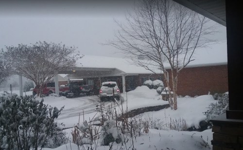 A snowy scene outside a house with a covered porch, cars in the driveway, and snow-covered bushes.