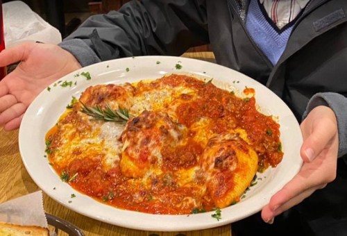 A person holds a large plate of cheesy ravioli topped with marinara sauce and garnished with a sprig of rosemary.