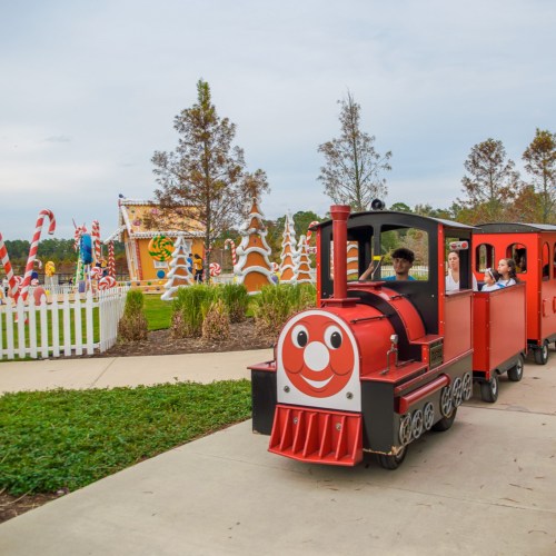 A colorful train with smiling faces carries children through a festive outdoor area with trees and decorations.
