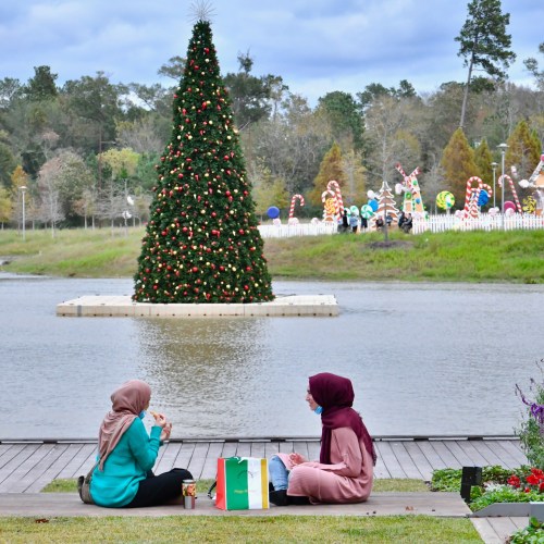 Two women sit by a lake, enjoying snacks, with a decorated Christmas tree and festive displays in the background.