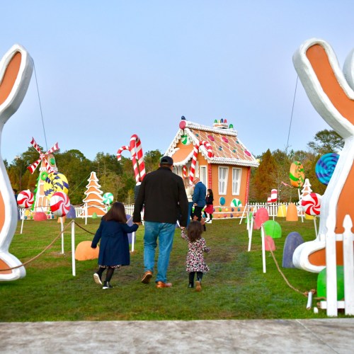 A family walks through a whimsical holiday display with giant candy and a gingerbread house in a festive outdoor setting.