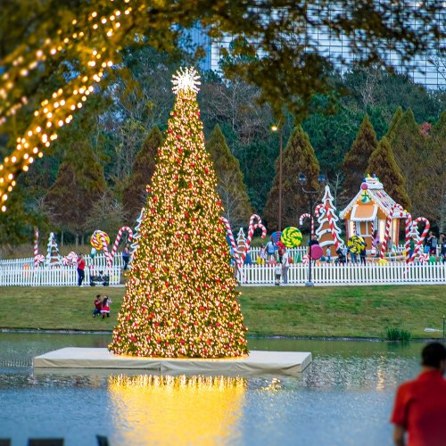 A brightly lit Christmas tree stands by a pond, surrounded by festive decorations and candy canes in the background.