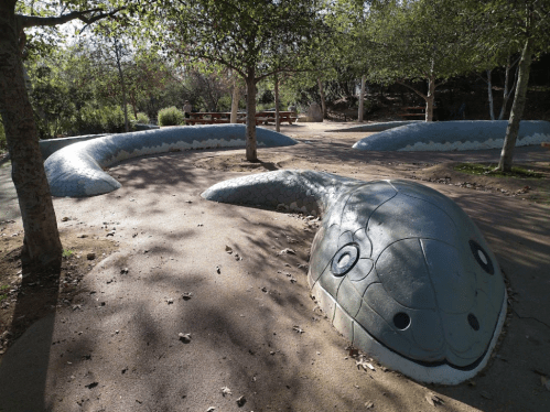 A large, mosaic snake sculpture winds through a park, surrounded by trees and a sandy path.