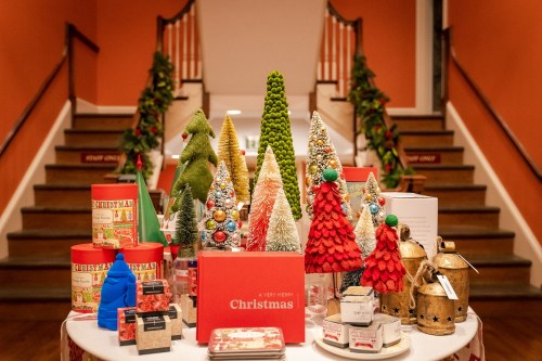A festive display of colorful Christmas trees and decorations on a table, with a staircase adorned with garlands in the background.