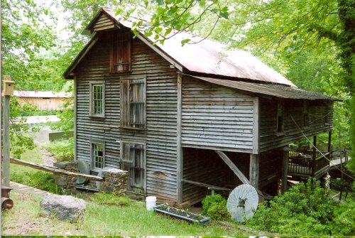 A weathered wooden building with a red roof, surrounded by greenery and stone features, showcasing rustic architecture.