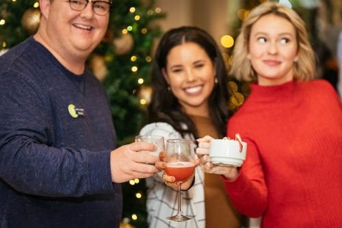 Three people celebrate with drinks in front of a decorated Christmas tree, smiling and enjoying the festive atmosphere.