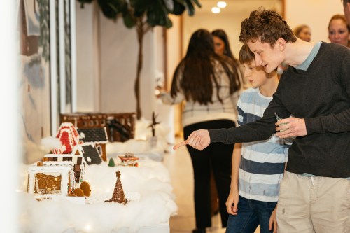 Two boys admire a display of gingerbread houses during a festive event, with people mingling in the background.