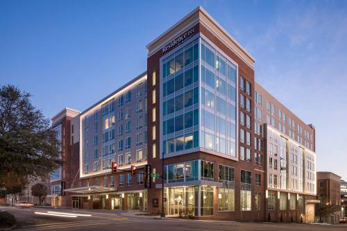 Modern multi-story hotel building with large windows, illuminated at dusk, located at a city intersection.