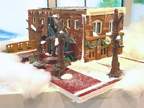 A festive gingerbread house with snowy decorations, surrounded by trees and a white, fluffy landscape.