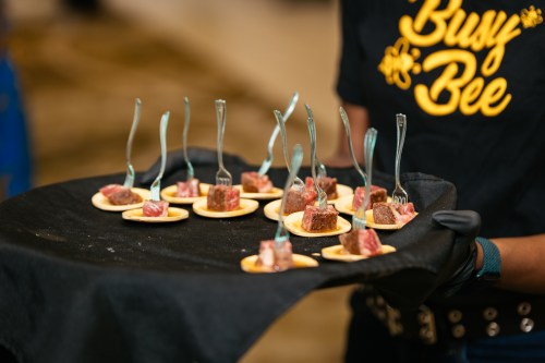 A server holds a tray of bite-sized meat appetizers, each with a small fork, at a catered event.