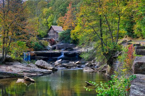 A scenic view of a historic mill by a tranquil stream, surrounded by colorful autumn foliage and rocky terrain.