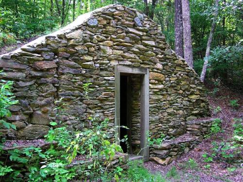 A weathered stone structure with a doorway, surrounded by lush greenery in a forested area.