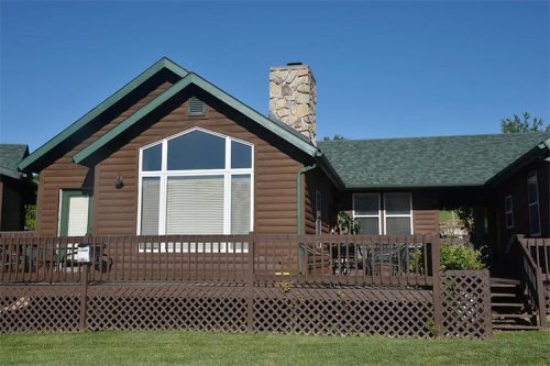 A brown log cabin with a stone chimney, large front windows, and a wooden deck surrounded by green grass.