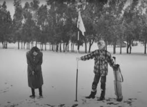 Two people in winter clothing stand on a snowy golf course, one holding a flagpole and the other looking down.