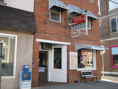 A brick building featuring "The Korner Cafe" sign, with a Coca-Cola logo and a small newspaper stand outside.