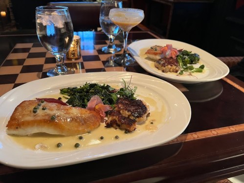 Two elegant plates of fish with sides, accompanied by glasses of water and cocktails on a checkered table.