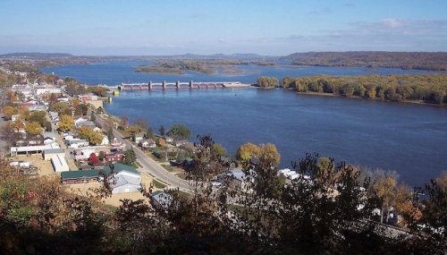 A scenic view of a river with a dam, surrounded by trees and a small town along the shore under a clear blue sky.