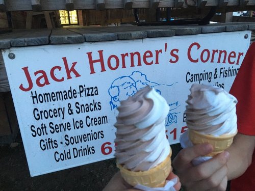 Two ice cream cones with swirled soft serve in front of a sign for "Jack Horner's Corner" offering snacks and camping supplies.