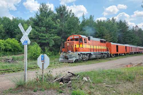 A bright orange train with a passenger car stops near a railroad crossing in a lush green landscape under a blue sky.