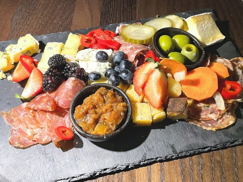 A charcuterie board featuring various cheeses, meats, fruits, pickles, and a bowl of chutney on a slate platter.