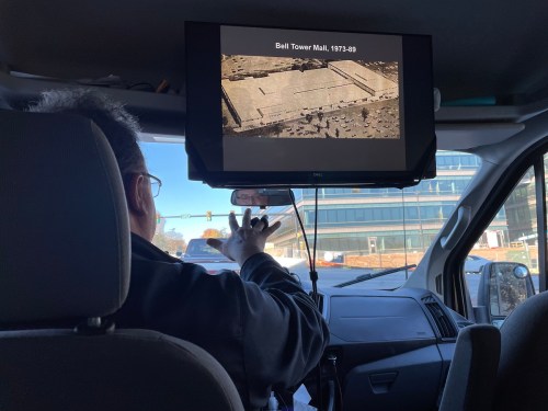 A person in a vehicle points at a screen displaying an image of Bell Tower Mall from 1973-49.