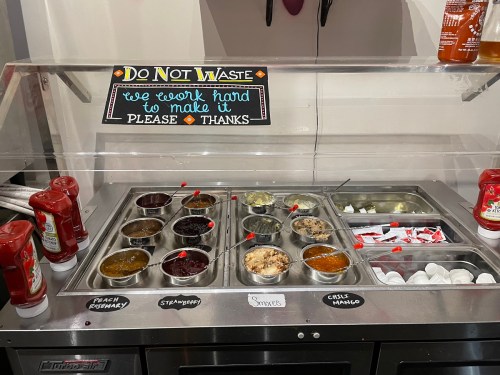 Condiment station with various sauces and toppings, labeled with signs urging not to waste. Ketchup and packets visible.