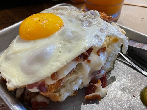 A close-up of a sandwich with bacon, melted cheese, and a sunny-side-up egg on top, served on a metal tray.