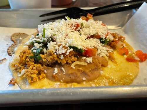 A close-up of a taco topped with refried beans, cheese, tomatoes, and herbs on a metal tray.