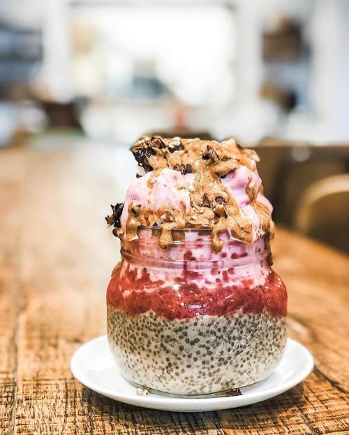A layered dessert in a jar with chia seeds, raspberry sauce, whipped cream, and chocolate drizzle on a wooden table.