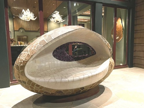 A modern, oval-shaped bench made of stone and tile, set against a backdrop of large windows and artistic decor.