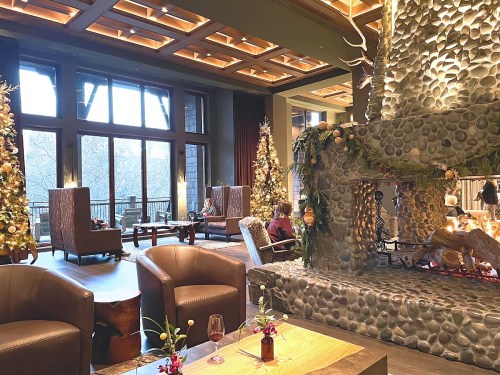 Cozy lodge interior with stone fireplace, festive Christmas trees, and comfortable seating by large windows.