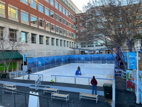 An outdoor ice skating rink surrounded by buildings, with a few people and benches nearby.