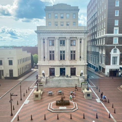 Historic building with columns, surrounded by a plaza and modern buildings, under a clear blue sky.