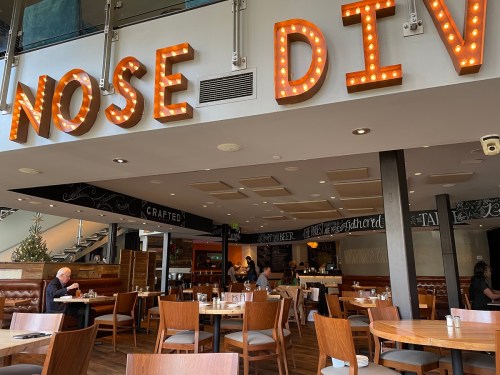 Interior of a restaurant named "Nose Dive," featuring wooden tables, chairs, and bright marquee-style signage.