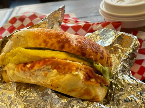 A toasted sandwich wrapped in foil, filled with egg, bacon, and avocado, with a side of takeout containers.