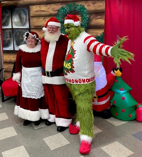 Santa, Mrs. Claus, and the Grinch pose together in a festive setting with holiday decorations.