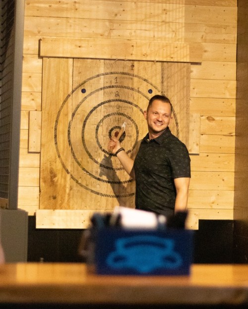 A man smiles while pointing at a wooden target board with concentric circles in a casual setting.