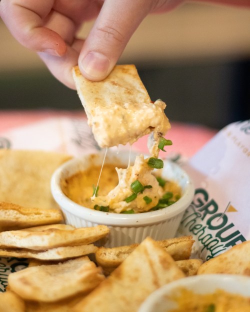 A hand dips a crispy chip into a creamy dip, with melted cheese and green onions visible. Surrounding chips are scattered.