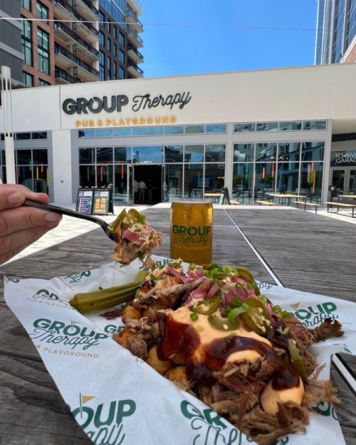 A hand holding a fork with a plate of pulled pork and toppings in front of a pub and playground with a drink.