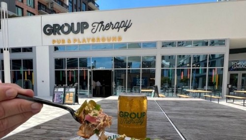 A hand holding a fork with food in front of the Group Therapy Pub & Playground, featuring a drink beside it.