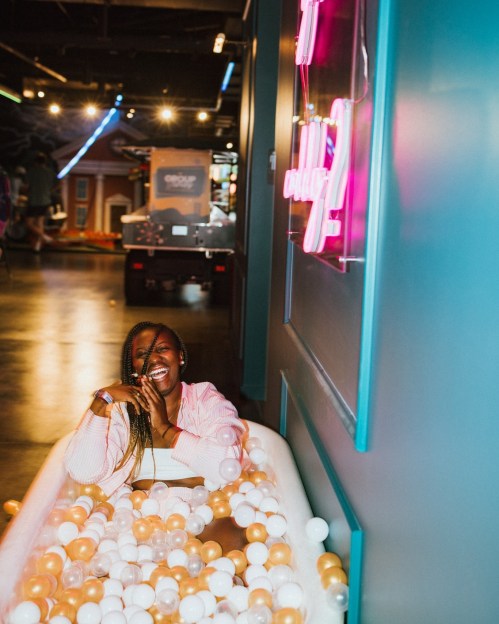 A person sits in a ball pit, smiling and wearing a pink outfit, with colorful lights and a neon sign in the background.