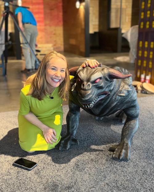 A woman in a green dress smiles while posing next to a large, menacing-looking creature on a carpeted floor.