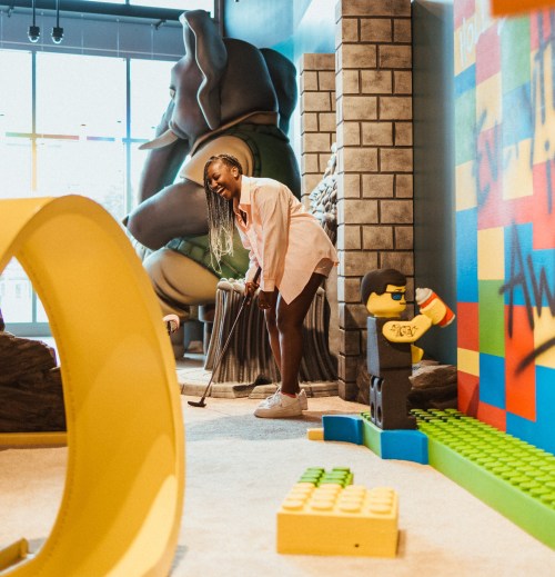 A person plays mini-golf in a colorful indoor space with large sculptures and playful decor.