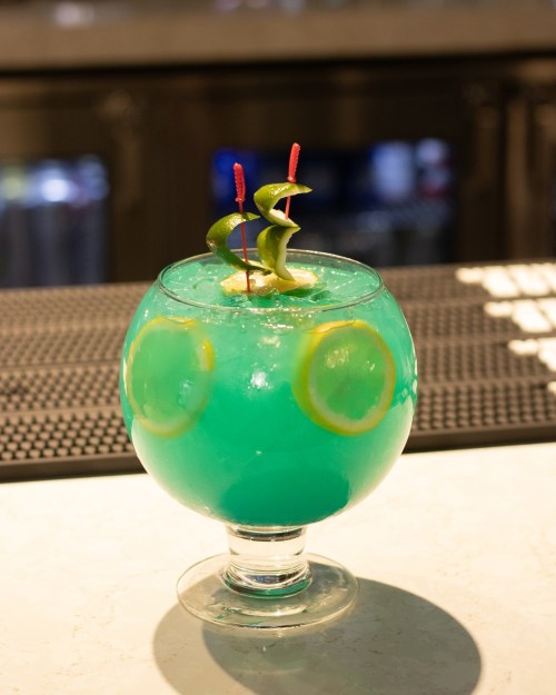 A vibrant blue cocktail in a round glass, garnished with lemon slices and decorative green twists.