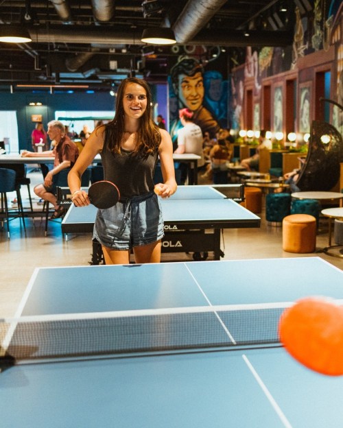 A young woman plays table tennis in a vibrant, lively venue with colorful murals and people in the background.
